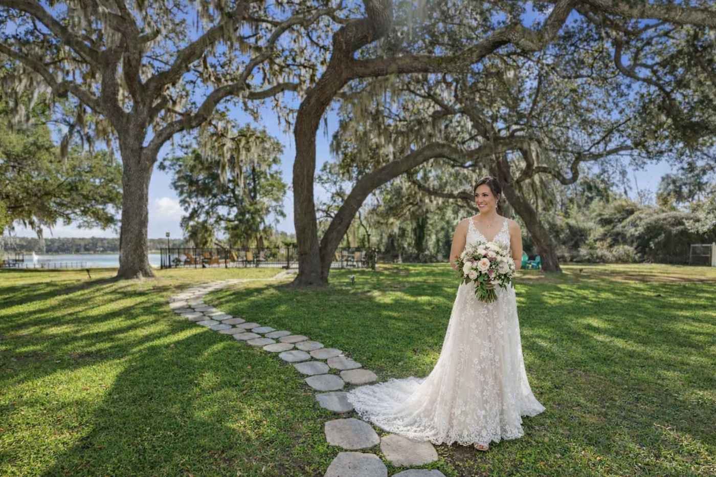 A bride holding her bouquet on the stone path that winds through the lawn under the live oaks toward the lake